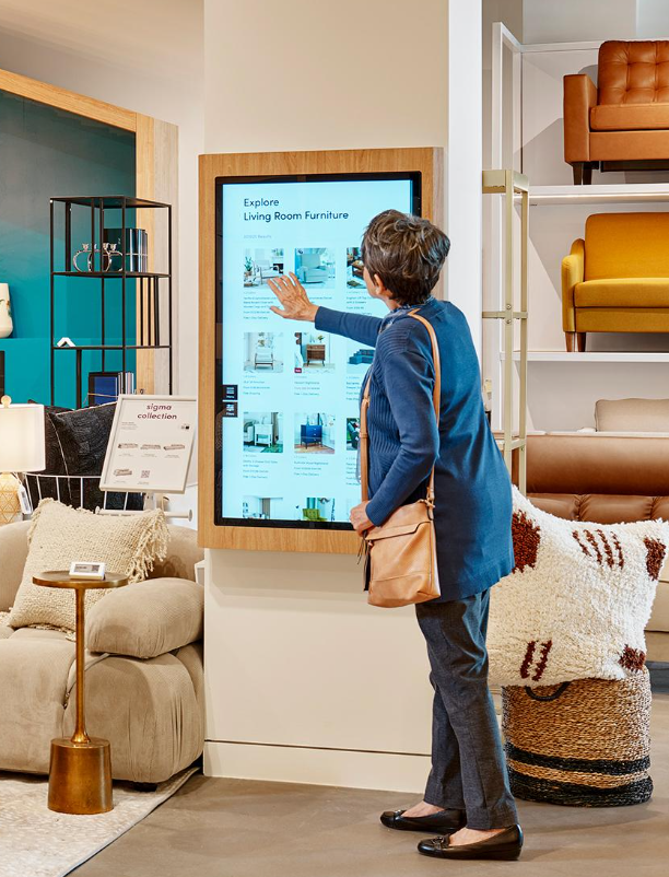 Klant gebruikt een interactief touchscreen in een meubelwinkel om meubels, productinformatie en verschillende uitvoeringen te bekijken.